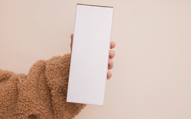 Woman hands in a cozy sherpa sweatshirt holds white cardboard box on a beige background图片下载