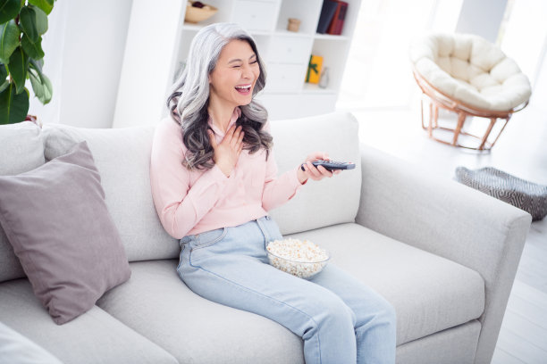 照片：一位快乐、愉快的漂亮老妇人在室内家中看电视喜剧，吃着爆米花，手里拿着遥控器。图片下载