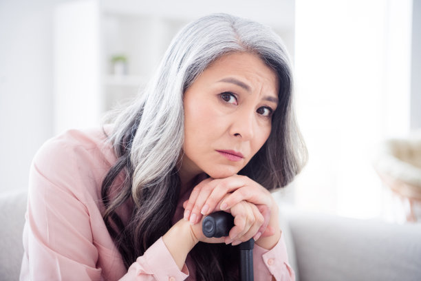 照片：一位不开心、悲伤的老妇人独自在家里的客厅里，手托着下巴，拄着拐杖。图片下载