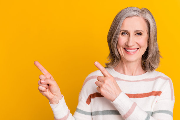一位穿着白色毛衣、面带微笑、手指指向黄色背景空白处的迷人退休女士的甜美照片图片下载