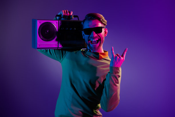 照片：一个时髦快乐的年轻男子，心情很好，竖起摇滚手势，手持录音机，背景是霓虹灯图片下载