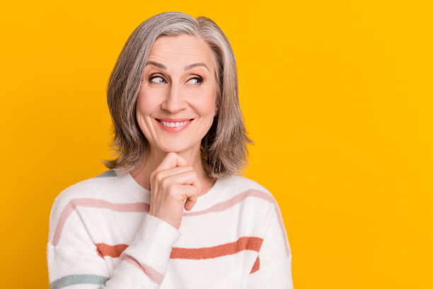 照片：一位乐观的灰发老妇人，手托下巴，望向远方，身穿毛衣，背景为黄色。图片下载