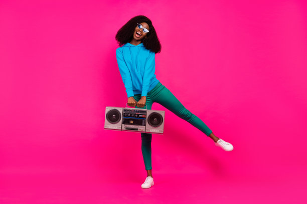 身穿蓝色运动衫的漂亮酷炫黑皮肤女子，在粉色背景前对着音响微笑跳舞图片下载
