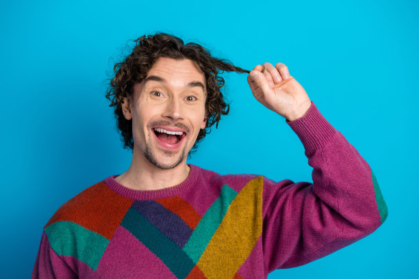 快乐的年轻男子穿着色彩鲜艳的菱格毛衣，在明亮的蓝色背景前对着镜头露出灿烂的笑容，俏皮地拨弄着他卷曲的头发，这是一张充满活力的时尚生活方式图片图片下载