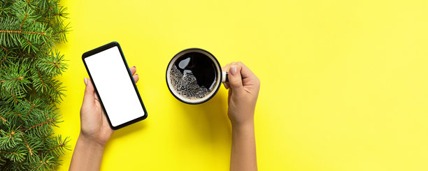 俯视视角，女性双手拿着黑色手机，手机屏幕为白色空白，旁边放着一杯咖啡。背景为黄色，采用平面布局，留有复制空间。图片下载
