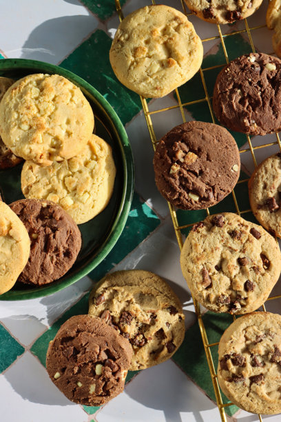 绿色盘子和金属冷却架上巧克力碎曲奇饼干圆的特写图片，节日三倍巧克力碎、牛奶巧克力碎和白巧克力碎饼干，白色和绿色格子图案背景，俯视角度图片下载