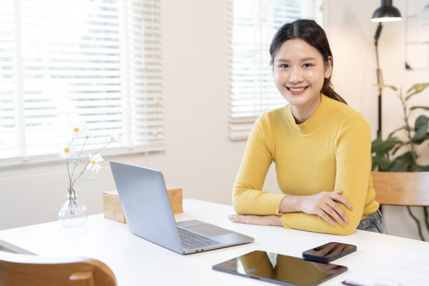 一位年轻的亚洲女性正在现代家庭办公室或工作空间使用笔记本电脑和数字设备工作，自信地对着镜头微笑。图片下载
