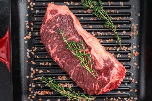 烤盘上的生西冷牛排，撒上百里香和盐，背景为深色石板，展现出牛肉的雪花纹理和质朴的烹饪风格。图片下载