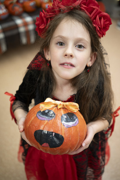 一名有发育障碍的女孩身穿吸血鬼服装，头戴花环，在万圣节背景下抱着一个装饰过的小南瓜的竖幅照片。图片下载