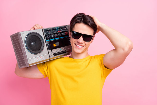 穿着黄色T恤、戴着墨镜的酷炫自信的年轻男子，微笑着听着收音机，背景是孤立的粉色。图片下载