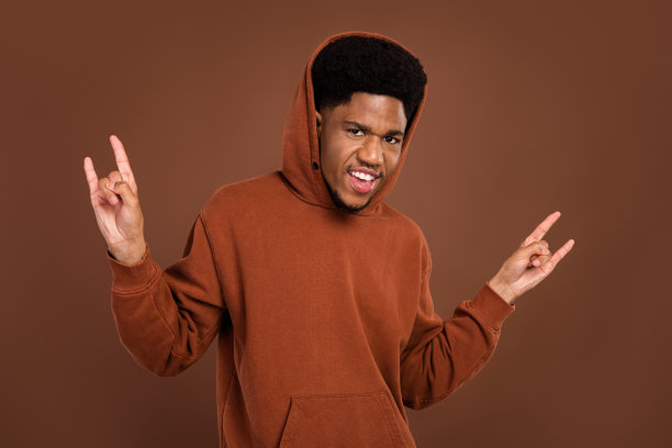 身穿运动服的酷炫棕色头发千禧一代男性，打着摇滚手势，在棕色背景中展示发型图片下载