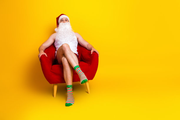 幽默的圣诞老人穿着条纹袜子，留着白胡子，在家里的红色椅子上放松，背景是黄色的，他微笑着，这是圣诞假期的生活方式照片。图片下载