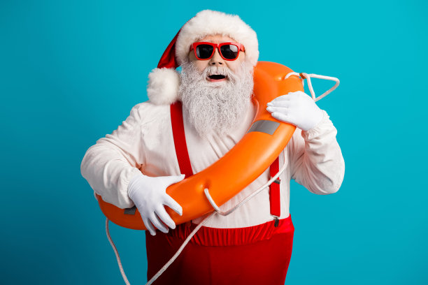 身穿红色吊带和白色套装的圣诞老人，戴着救生圈，在明亮的蓝色背景前摆姿势，用于假日季的航海主题素材照片图片下载