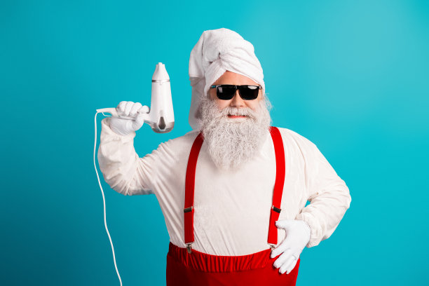 圣诞老人穿着白色西装，系着红色吊带，头裹白色毛巾头饰，在蓝色背景前用吹风机吹头发，打造节日氛围的素材照片图片下载