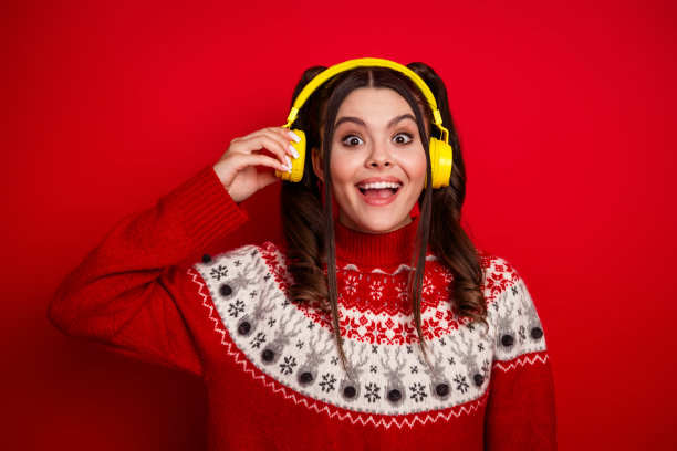 一位戴着亮黄色耳机的年轻女子，身穿带有北欧图案的节日红色毛衣，在鲜红的背景前摆姿势，作为圣诞节素材照片图片下载
