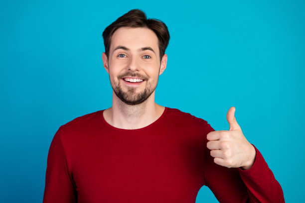 一名穿着红色毛衣的年轻男子，在蓝色背景前对着镜头竖起大拇指，笑容满面，适合用作广告和生活方式图片图片下载