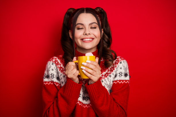 一位穿着红色毛衣的快乐年轻女子，手中拿着装有棉花糖的马克杯，背景为红色，捕捉了节日冬季的温暖，适合作为假日季的图片素材图片下载