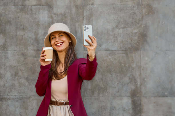 时尚女性在城市墙壁背景前手持咖啡杯自拍，用于时尚图库照片图片下载