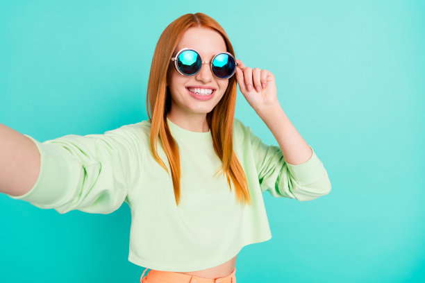 一位红发女孩戴着太阳镜，微笑着自拍，背景是充满活力的蓝绿色，照片肖像图片下载