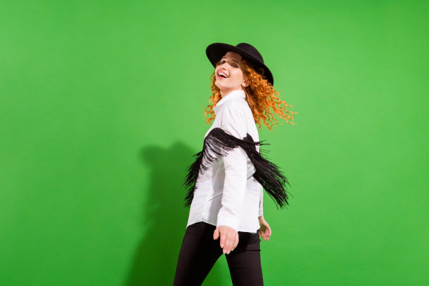 千禧一代女士的搞笑橙色发型个人资料照片，她身穿衬衫、裤子和帽子，站着看向镜头，孤立在绿色背景上图片下载