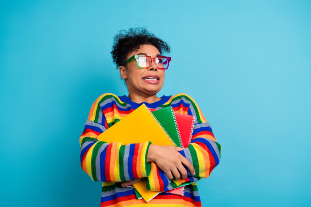 年轻的混血女性穿着色彩鲜艳的条纹毛衣，手持笔记本，在明亮的蓝色背景前微笑，这是一幅充满活力的休闲时尚生活方式图片。图片下载