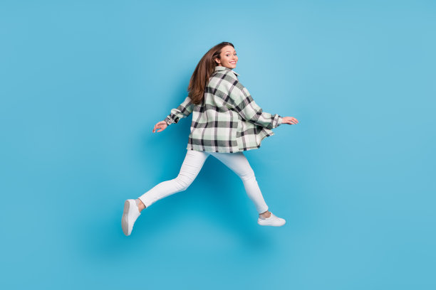 一名快乐友善的年轻女子全身侧面照，她正跳起来奔跑，身处蓝色背景的空白空间中图片下载