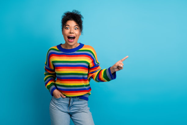 年轻女子穿着色彩条纹毛衣，对着蓝色背景微笑并指向前方，适用于生活方式时尚广告和动态品牌视觉设计图片下载