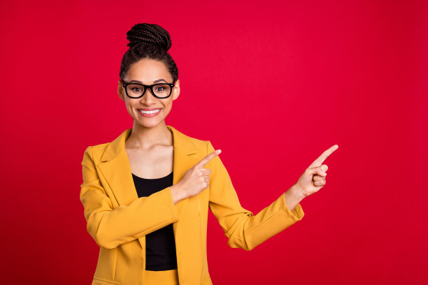 戴着眼镜、穿着黄色开衫、露出牙齿的女老师，在鲜艳的红色背景前，指向空白空间。图片下载