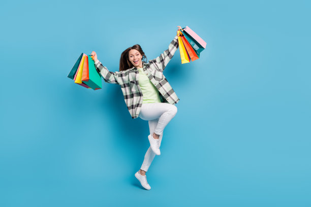 一位年轻女子举着购物袋，快乐地跳起来庆祝胜利，背景是蓝色的，她看起来很积极，这是这张照片的全尺寸图。图片下载