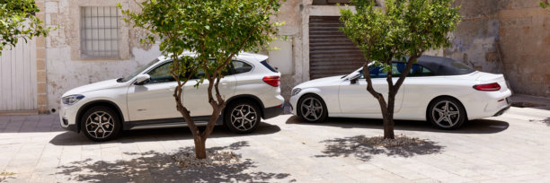 两辆白色豪华汽车，一辆宝马X1 SUV和一辆梅赛德斯-奔驰C级敞篷车，停在石头庭院的柠檬树旁。这两辆车与乡村建筑相得益彰。2022年6月26日，意大利，莫诺波利。图片下载