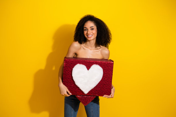 一位卷发年轻女子手持红色心形礼品盒，置身于明亮的黄色背景前，用于时尚别致的生活方式和时尚宣传图片图片下载