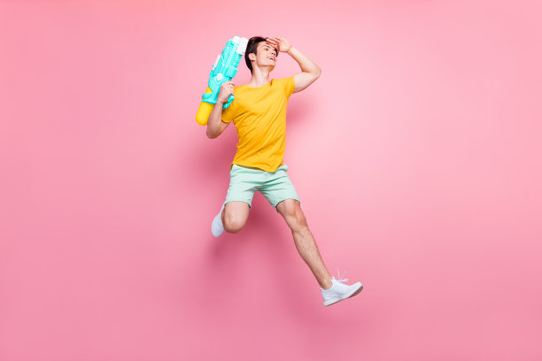 身穿休闲装的年轻男子，面带快乐积极的笑容，向前跳跃，手持水枪，在粉色背景下拍摄的全尺寸照片图片下载