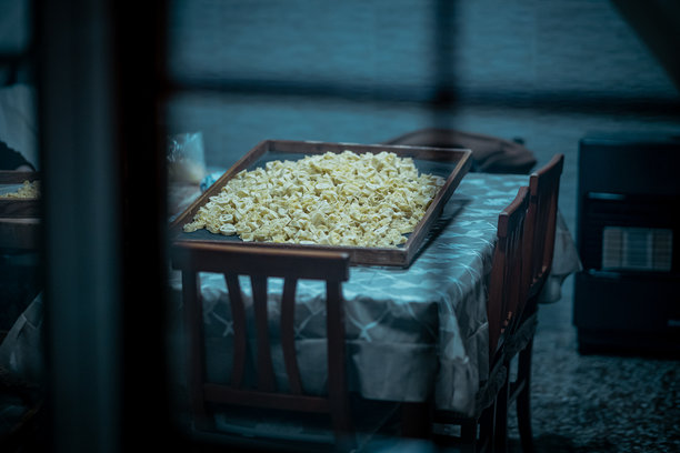 一张充满氛围、偷窥视角的照片，展示了桌上晾晒的新鲜手工小饺子（如意式馄饨），透过模糊的窗户拍摄，突显了温馨的意大利厨房场景。图片下载