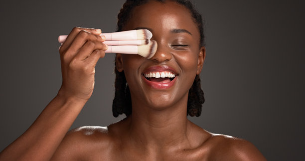 工作室里，一位快乐的黑人女性正在使用化妆刷和化妆品进行美妆，包括化妆、涂抹和美容产品。背景为深色，展示了美容学、空间及使用工具的个人。图片下载