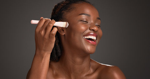 黑人女性，在工作室里带着化妆刷开心地笑着，背景是深色空间和粉底。非洲女孩，快乐地拿着化妆工具，对着产品兴奋地涂抹遮瑕膏和粉底，打造精致肌肤。图片下载