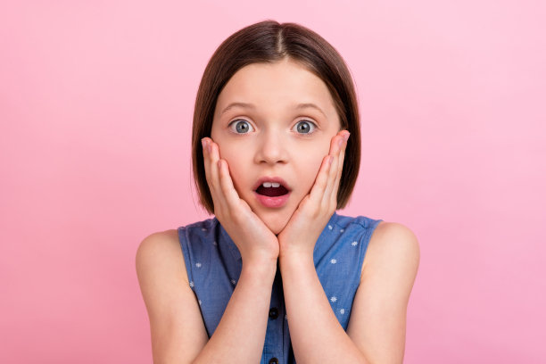 照片肖像，一名女学生惊讶地盯着看，双手触碰脸颊，似乎遇到了麻烦，背景是柔和的粉色。图片下载