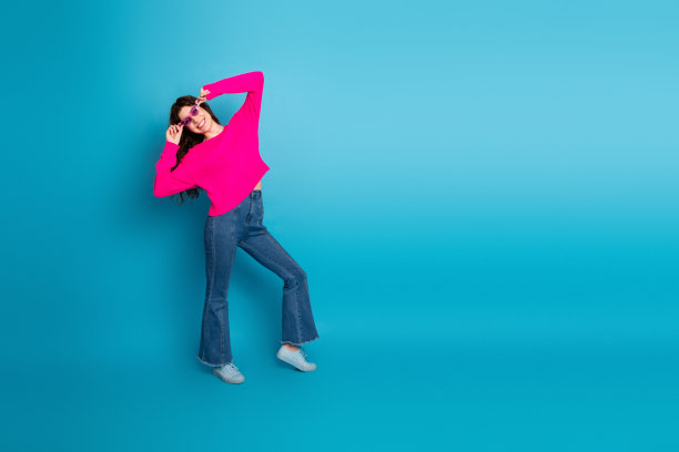 年轻女子身穿粉色针织毛衣，在蓝色工作室背景前快乐地摆姿势，用于生活时尚图片图片下载