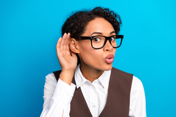 戴眼镜的年轻女子穿着白色衬衫和棕色马甲，好奇地倾听，背景为蓝色，非常适合商务时尚生活方式的图库照片图片下载