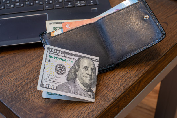 笔记本电脑旁的木桌上放着一个黑色皮质钱包，里面有一张百元美元钞票。 财务，个人预算，在线支付。真实照片图片下载