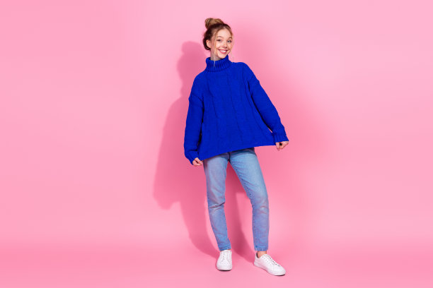 年轻女子穿着蓝色针织毛衣，在粉色背景前摆出俏皮姿势，打造时尚生活方式图片图片下载