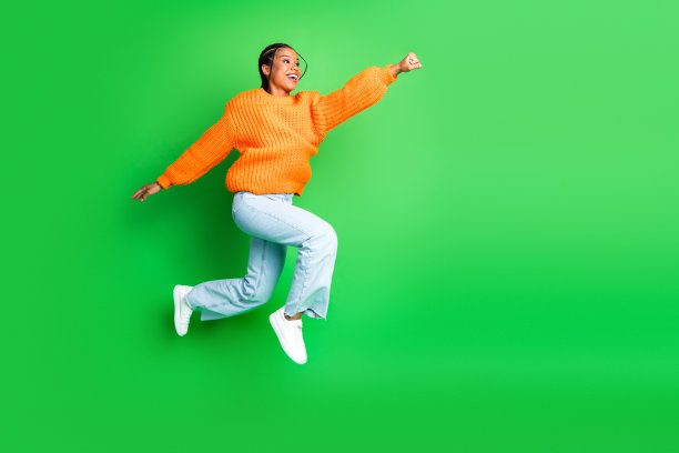 身穿橙色毛衣的年轻女子在绿色背景前跳跃，展现活力、快乐与时尚，适用于生活方式、健身及宣传素材。图片下载
