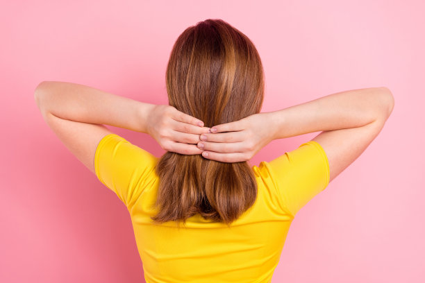 粉色背景下，年轻女学生的背部、后侧照片，她正在给自己做发型，使用护发乳，面部模糊处理，孤立显示。图片下载