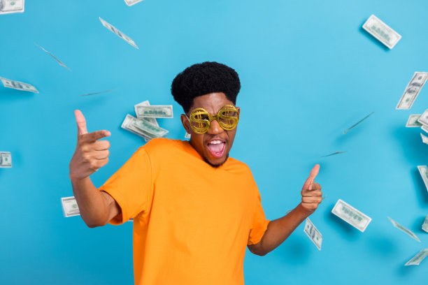 照片：一个有吸引力的年轻非裔美国男性，戴着眼镜，心情愉快，身上掉落着钱，背景是蓝色的，孤立的。图片下载