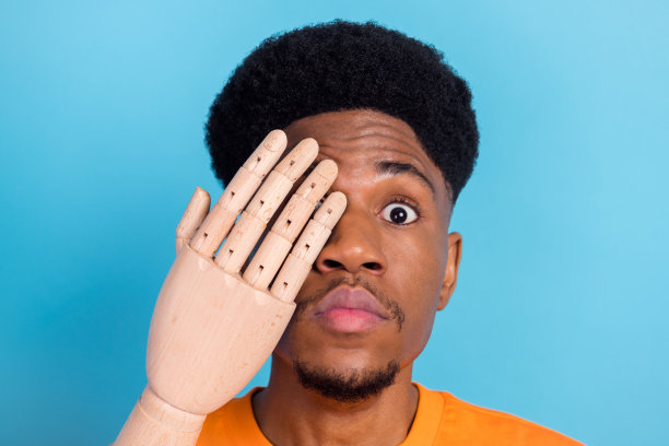 照片：惊讶的年轻黑皮肤男子用木制手遮住一只眼睛和脸，背景是柔和的蓝色图片下载