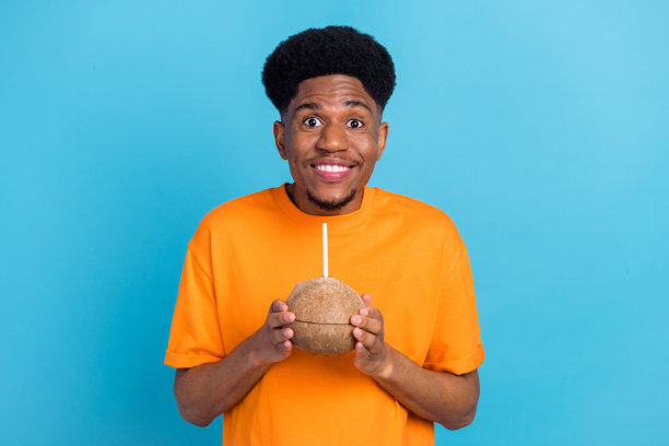照片：一位惊讶而快乐的非裔美国年轻男子，手持椰子鸡尾酒，背景为蓝色。图片下载