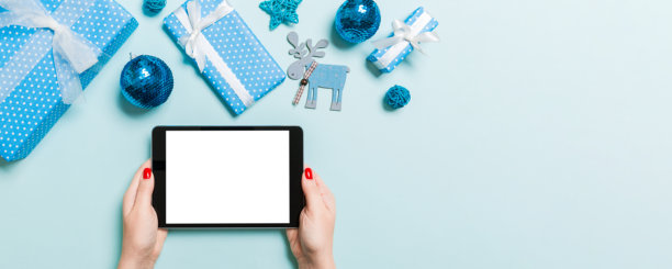 俯视角度，一名女子双手拿着平板电脑，背景是蓝色的圣诞装饰品。新年节日概念，样机。图片下载
