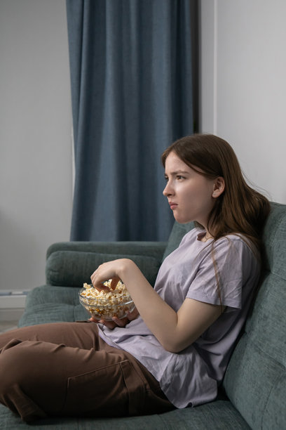 一个厌倦的少女坐在客厅的绿色沙发上，看着无聊的电影或电视节目，用玻璃碗吃着爆米花，脸上流露出失望和冷漠的神情。竖直照片。图片下载