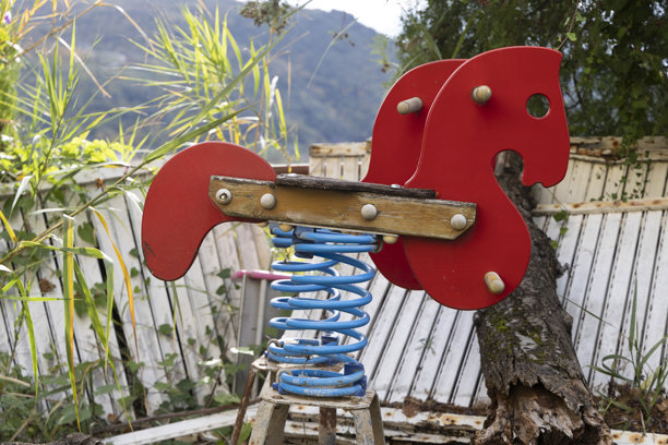 一张废弃的游乐场照片，在被忽视的户外区域有三匹生锈、破旧的红色弹簧马。图片下载