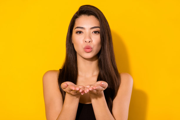 照片：一位迷人的年轻女子，脸上带着积极的表情，双手做出飞吻的动作，在黄色的背景下显得有些调皮，孤立拍摄。图片下载