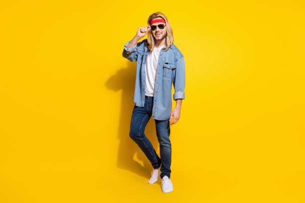 穿着牛仔衬衫、戴着墨镜、走在黄色背景前、面带微笑的滑稽帅气年轻男士照片图片下载